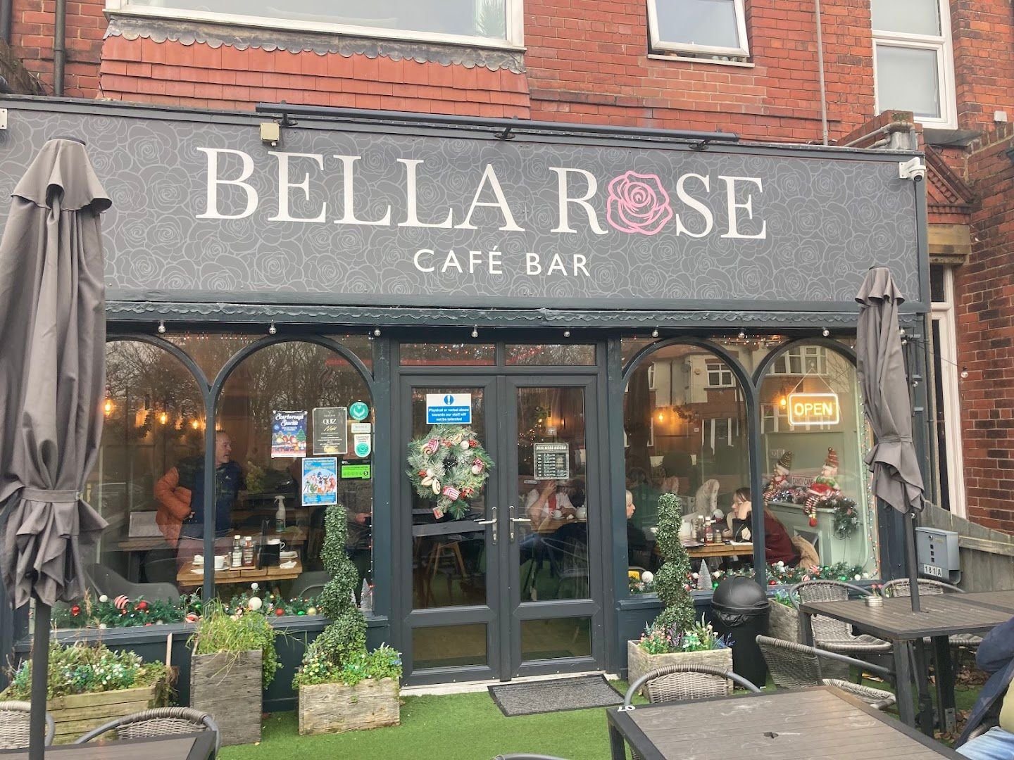 The Bella Rose shopfront: a dark grey sign reading Bella Rose Café Bar with a pink rose for the O, arched windows, and tables, planters and topiary on the terrace in front